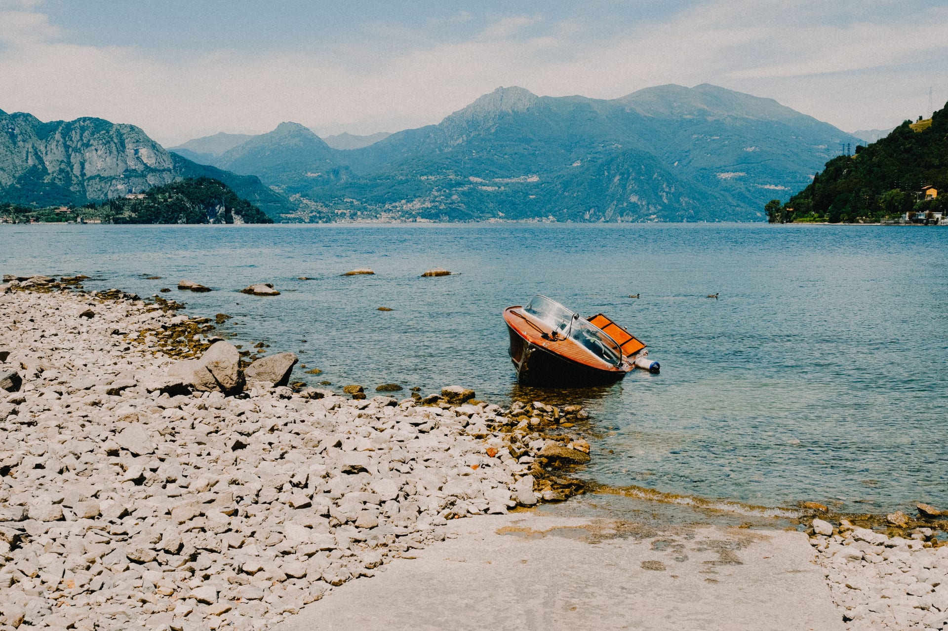 Riva di ciottoli e sassi sul lago, con una barca di legno e le montagne sullo sfondo