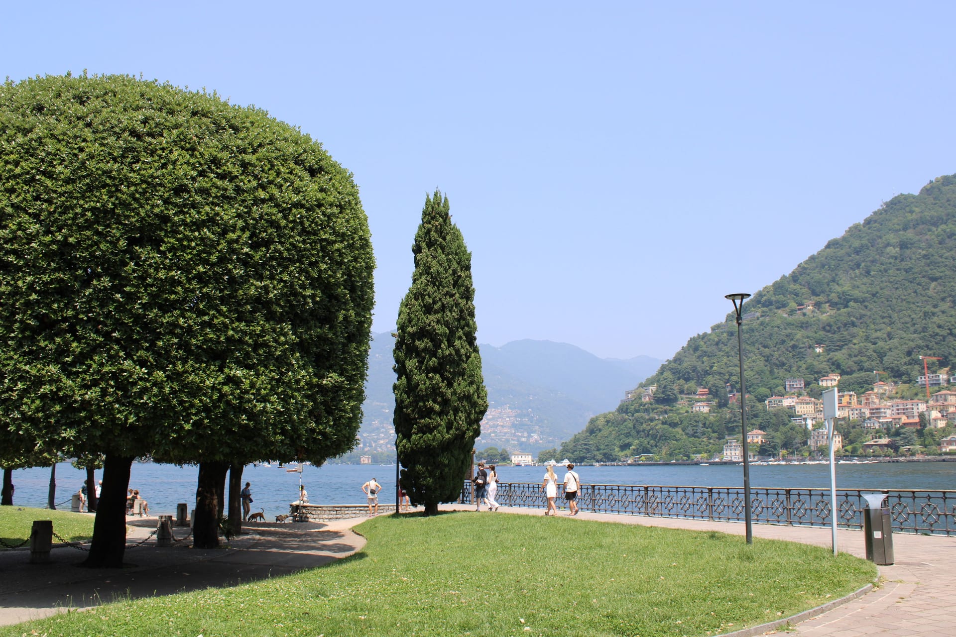 Prato alberato e passeggiata sul lungolago di Como, con un paese colorato sull'altra sponda