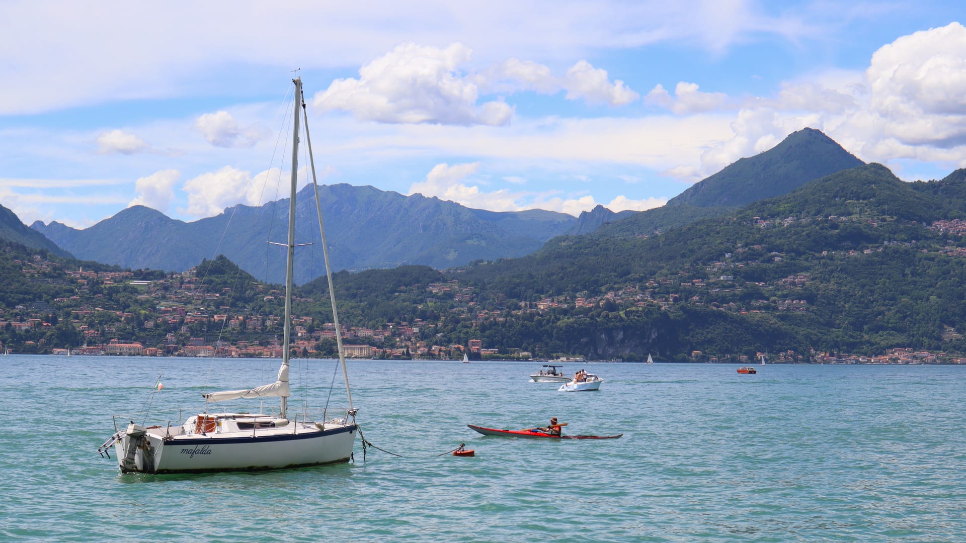 Barca a vela e kayak sul Lago di Como in una giornata estiva, con un borgo sullo sfondo