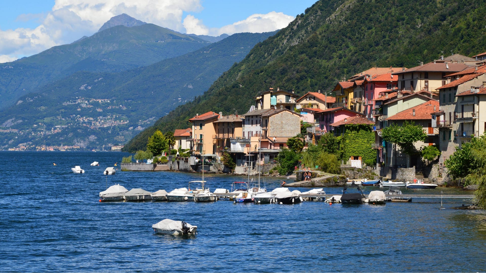 Barche ormeggiate davanti a un borgo colorato sulla riva del Lago di Como, con le montagne sullo sfondo