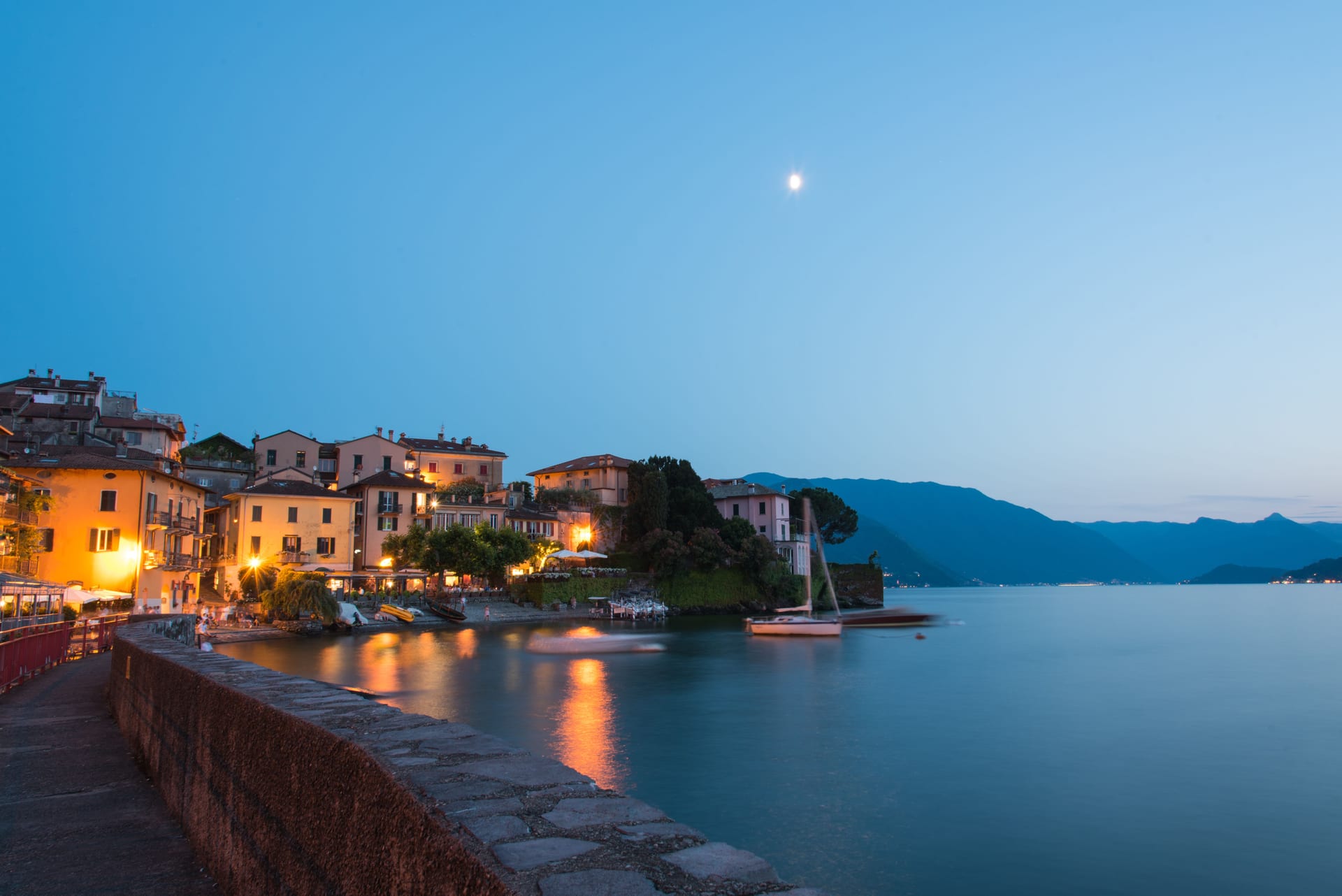 Un borgo del Lago di Como illuminato al crepuscolo, con le case colorate affacciate sull'acqua e la luna nel cielo