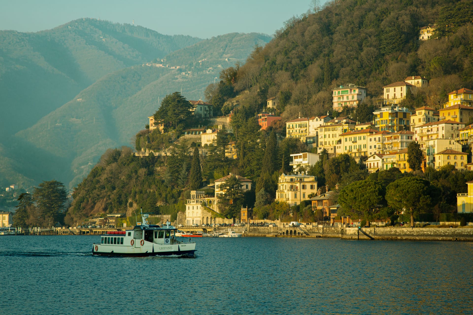 Un traghetto passeggeri naviga davanti a un borgo del Lago di Como con case colorate sulla collina