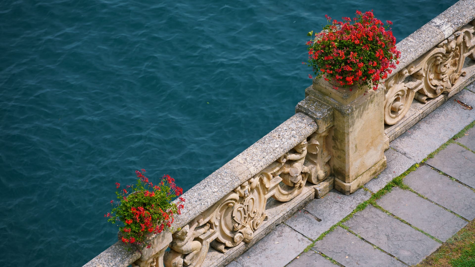 Balaustra in pietra con vasi di gerani rossi affacciata sulle acque del Lago di Como
