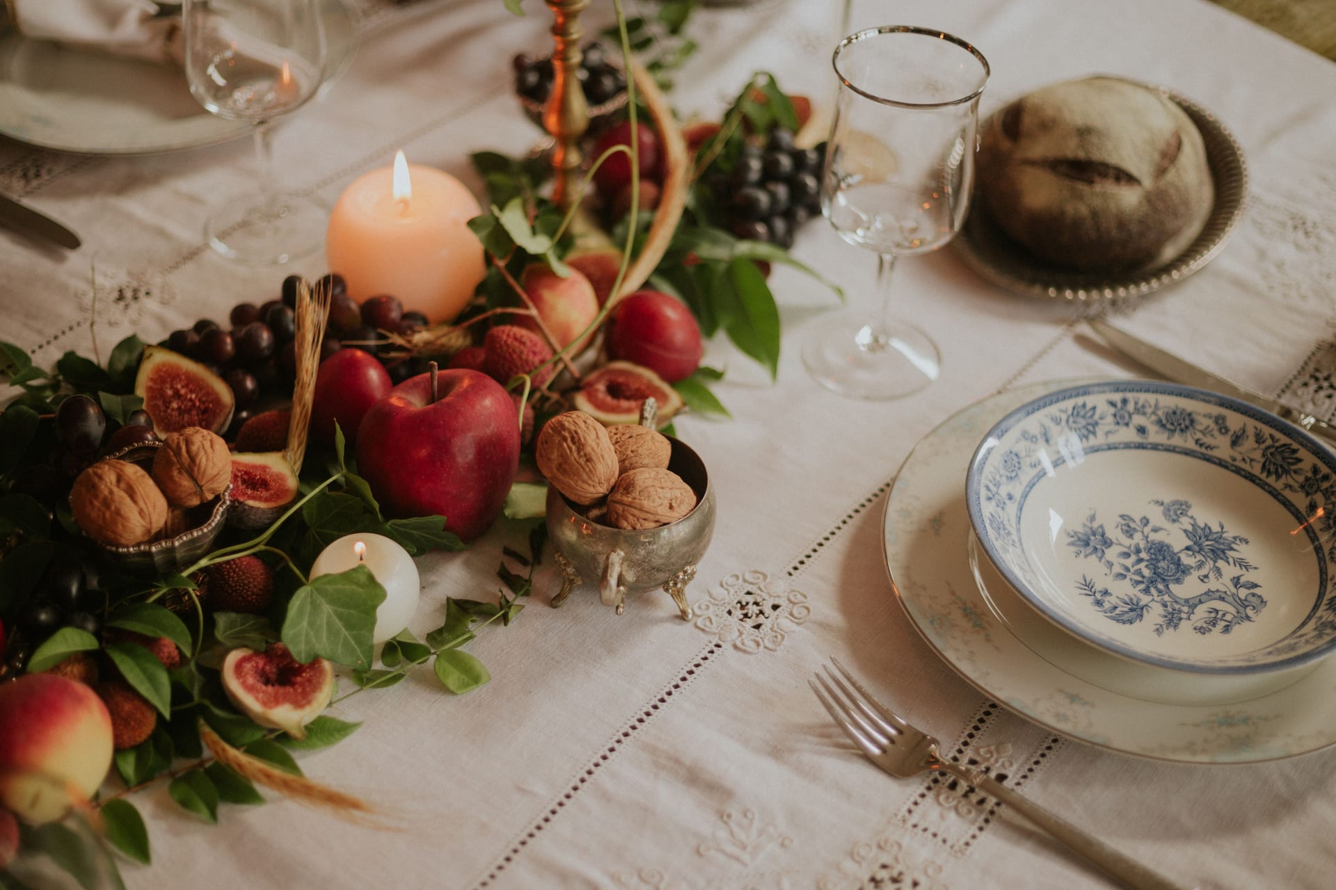Tavola apparecchiata con tovaglia di lino ricamata, ceramiche decorate, candele accese e frutta d'autunno: fichi, mele, uva e noci