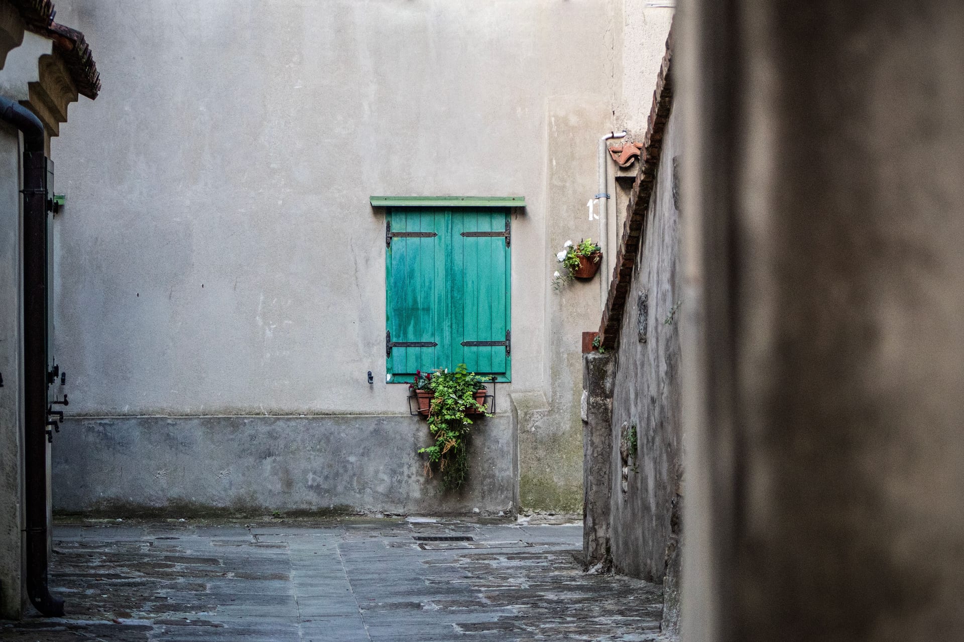 Vicolo acciottolato di un borgo della Tremezzina, con una persiana verde e vasi di gerani lungo il muro in pietra